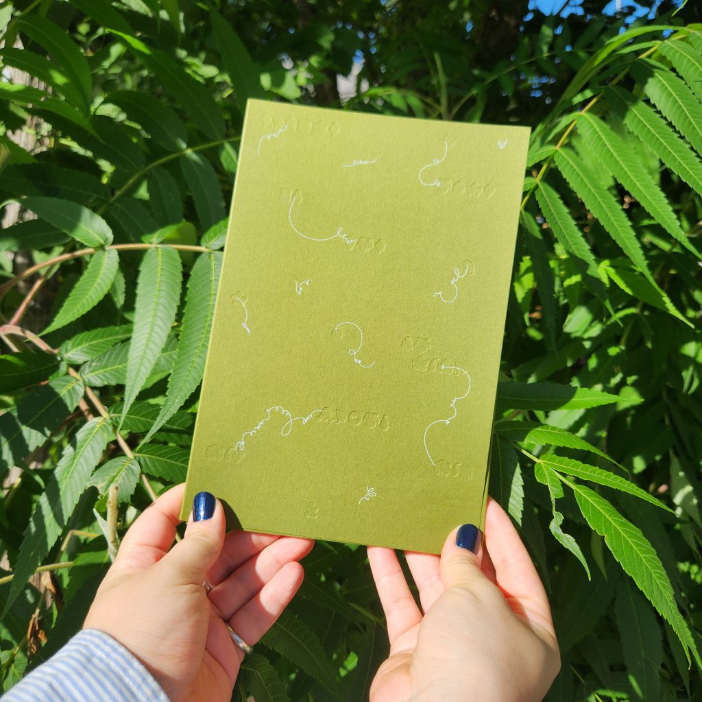 Two hands, with nails painted dark metallic blue, hold a pear-coloured book adorned with small embossed shapes and white squiggly lines. In the background is a leafy sumac shrub.