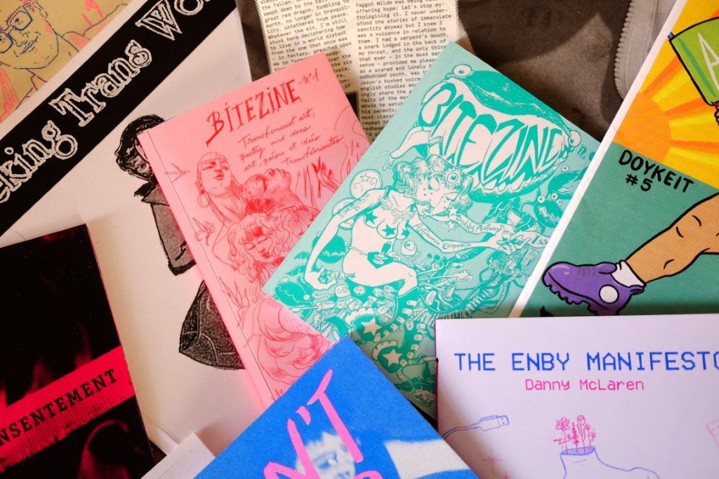 Bitezine, Vols. I & II, appear surrounded by an array of other colourful zines. Vol. I features a pink background with a red, line-drawing of various femme figures, and Vol. II features a turquoise background, with a darker turquoise illustration of a thin, three-breasted mermaid-type figure.