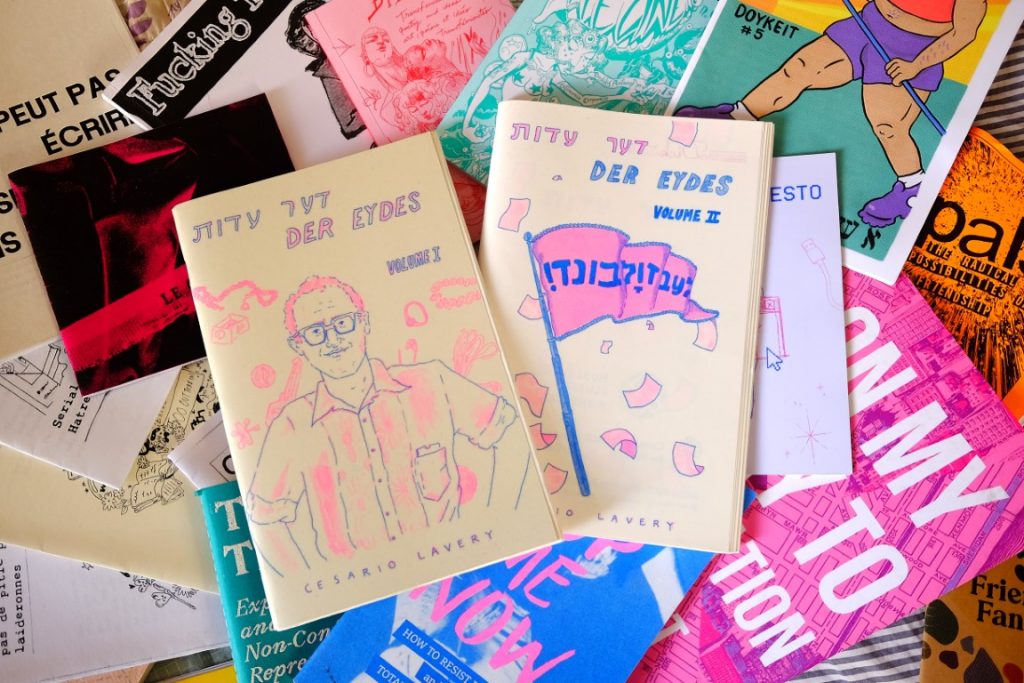 Der Eydes, Vols. I & II, lie on a scattered array of other colourful zines. Their covers are off-white with hot pink and blue illustrations, and the one on the left features a figure with short hair, thick-framed glasses, and a button-up shirt, while the one on the right features a pennant flag with Hebrew lettering.